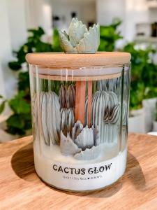 Świeca sojowa w szkle z drewnianym knotem i zielonymi kaktusami. Na drewnianej pokrywce dekoracyjny kaktus.