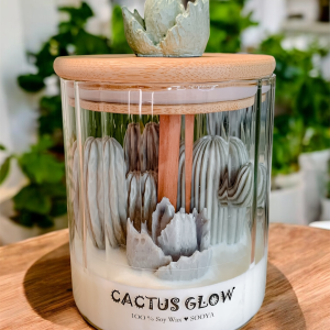 Świeca sojowa w szkle z drewnianym knotem i zielonymi kaktusami. Na drewnianej pokrywce dekoracyjny kaktus.