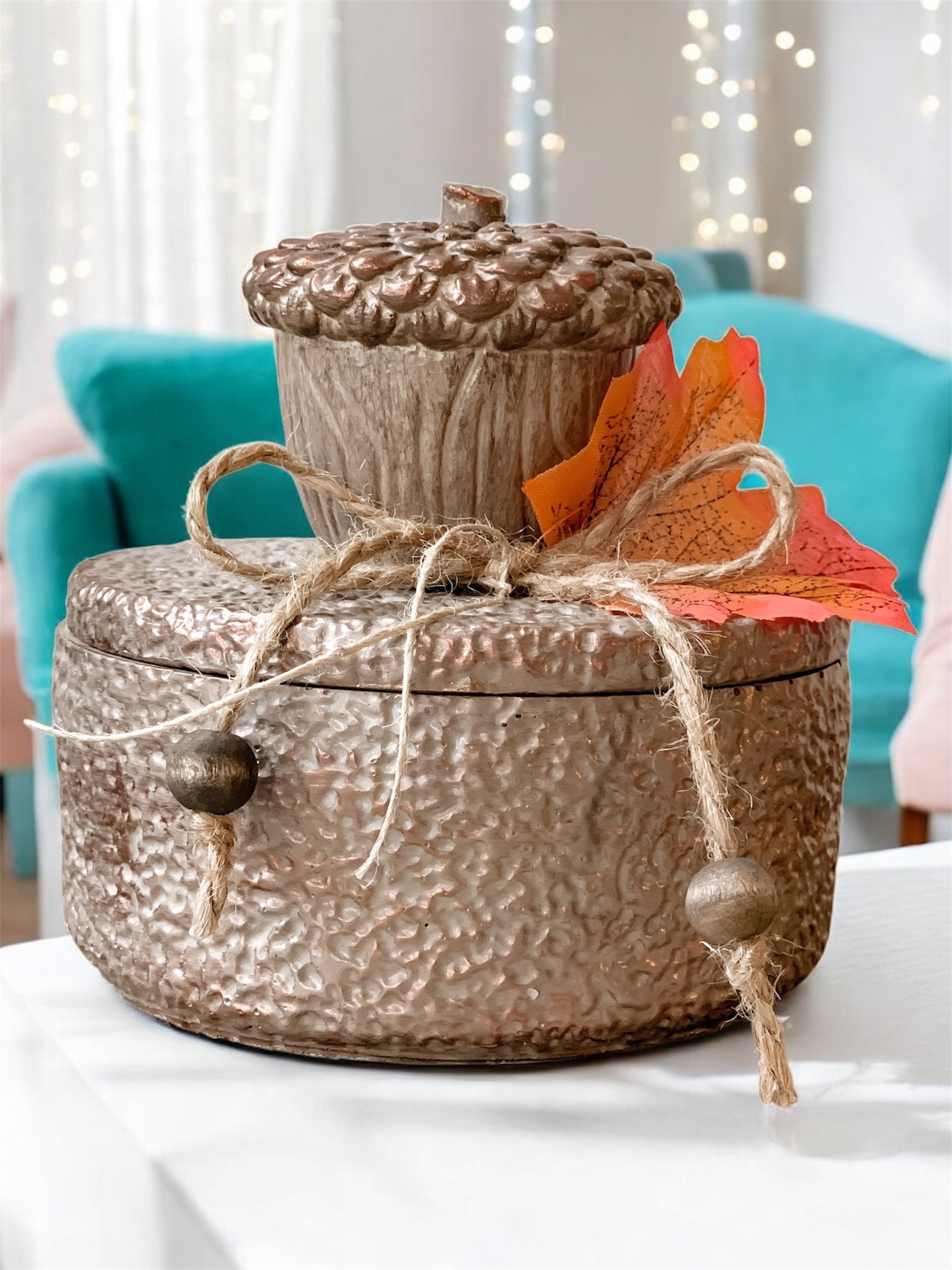 Jesienne Żołędzie świeca w brązowym pojemniku z pokrywką. Ozdobiona dużym brązowym żołędziem i jesiennym liściem, przywiązanym sznurkiem jutowym.