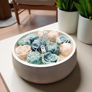 Świeca dekoracyjna Kwiatowy Ogród w okrągłym pastelowym pojemniku z gipsu, udekorowana kwiatami w odcieniach błękitu i ecru. Na jednym z kwiatów dekoracyjny motylek.